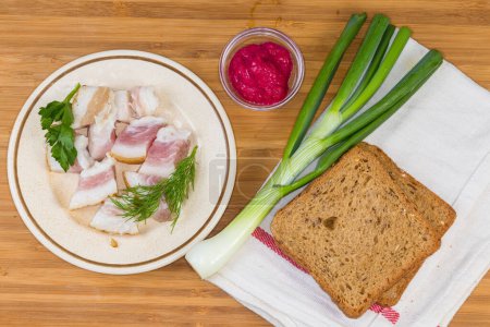 Small slices of the salted pork fatback on skin with meat layer on a saucer,  bread,  stem of green onion and horseradish sauce on a wooden cutting board, top view