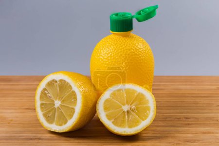 Concentrated lemon juice in yellow plastic container with open green dosing cap and two halves of fresh lemon on a wooden surface