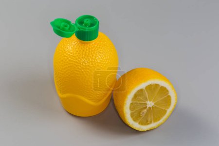 Concentrated lemon juice in yellow plastic container with open green dosing cap and half of fresh lemon on gray background