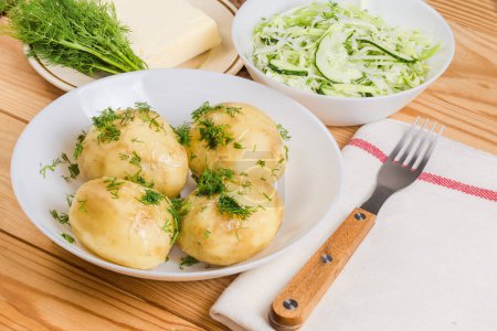Serving of the boiled whole young potatoes with butter and sprinkled with chopped fresh dill and green vegetable salad, butter piece and bunch of dill on a saucer on a rustic table, selective focus