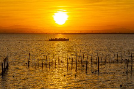 Photo for The boat with tourists and fishermen on a lake with fish nets and sunny path against the backdrop of an orange sunset - Royalty Free Image