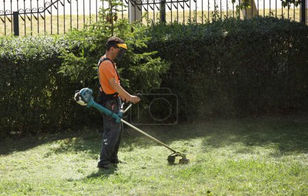 Foto de A worker mows grass with a mechanical trimmer. Improvement of a green lawn with a manual lawn mower with an internal combustion engine. Work with landscape design - Imagen libre de derechos