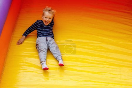 Téléchargez les photos : A hilarious little boy slides down at a high speed off a large, bright yellow trampoline. Child on the move, there is free space for text in the photo. - en image libre de droit