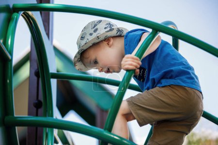 Téléchargez les photos : Heureux garçon de 3 ans grimpant et jouant à l'aire de jeux extérieure dans le parc de la ville. - en image libre de droit