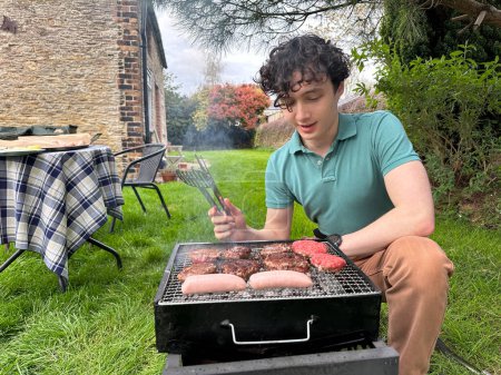 Foto de Joven haciendo barbacoa en el patio trasero, asando carne en una barbacoa portátil - Imagen libre de derechos