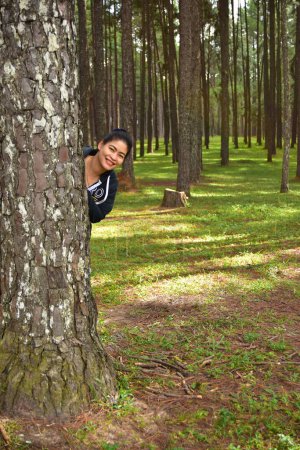 Téléchargez les photos : Souriant asiatique femme se cacher derrière un arbre dans pinède - en image libre de droit