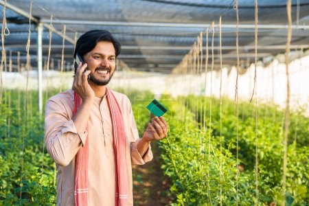 Téléchargez les photos : Young farmer at greenhouse talking on mobile phone call by holding green credit card - conept of online payment, customer service and agronomy. - en image libre de droit