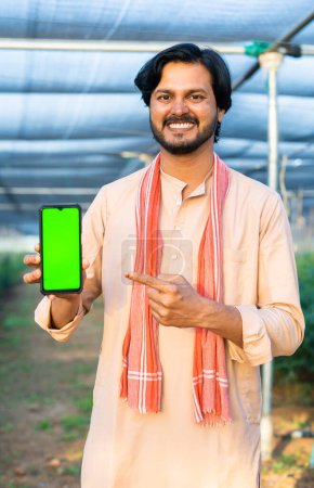 vertical shot of Happy smiling young farmer showing green screen mobile phone by pointing finger by looking camera at greenhouse - concept of agriculture promotion, advertisement and app promotion.