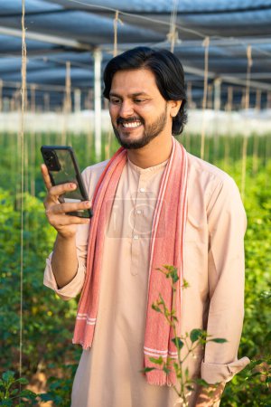 Téléchargez les photos : Vertical shot of Happy smiling young farmer busy using mobile phone at greenhouse - concept of technology, modern farming and agri business. - en image libre de droit