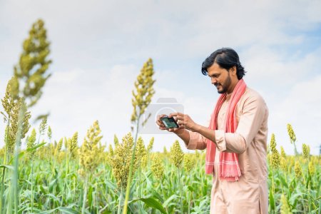Foto de Happy young farmer checking crop growth by taking photos on mobile phone app at farmland - concept of technology, modern farming and internet. - Imagen libre de derechos