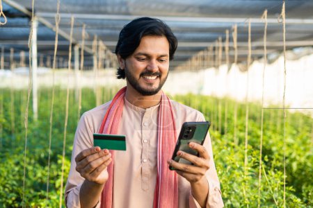 Happy Indian farmer with Green card using mobile phone at greenhouse - concept of online banking, application and technology.