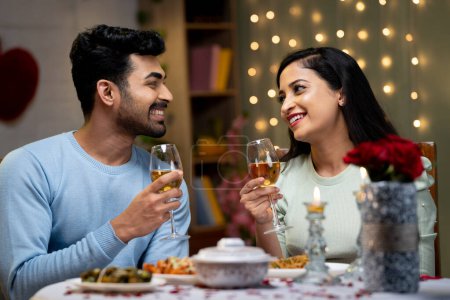 Happy smiling couple having drinks or wine by talking each other at candle light dinner at home - concept of romantic evening, dating and engagement plans