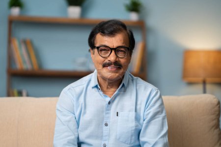 Photo for Happy indian senior man with eyeglasses by looking at camera while sitting on sofa at home - concept of successful, confident and satisfaction - Royalty Free Image