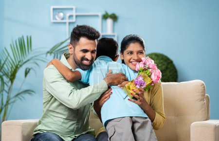 Téléchargez les photos : Enfant indien donnant des fleurs avec un cadeau aux parents en s'embrassant tout en étant assis sur le canapé à la maison concept de relation, célébration d'anniversaire et convivialité - en image libre de droit