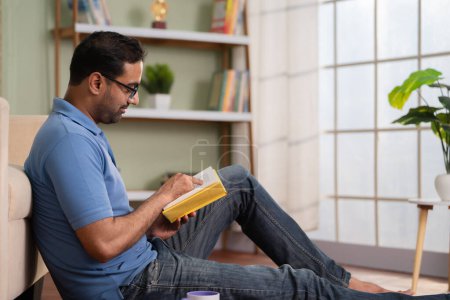 Téléchargez les photos : Homme indien avec des lunettes de lecture livre ou roman tout en étant assis sur le sol à la maison concept de passe-temps week-end, auto-amélioration et temps libre - en image libre de droit