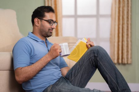 Téléchargez les photos : Coup de main de l'homme indien lisant le livre en buvant du café ou du thé tout en étant assis sur le sol à la maison concept de rafraîchissement, de connaissances et de passe-temps week-end. - en image libre de droit