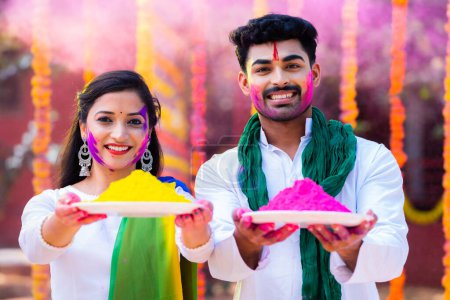Happy young colorful couple holding holi colour plate by looking at camera at flower decoration background - concept of holi festival, enjoyment and indian traditional.