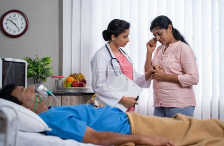 Médico indio que consuela a la esposa del paciente enfermo al dar confianza frente al marido dormido en la cama en la sala del hospital - concepto de estímulo, empoderamiento y apoyo médico