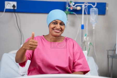 Glücklich lächelnde indische Brustkrebspatientin zeigt Daumen hoch, indem sie mit der Kamera auf das Krankenhausbett blickt - Konzept einer erfolgreichen Behandlung, inspirierende und positive Emotionen