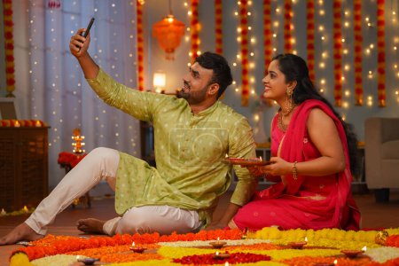 Téléchargez les photos : Heureux couples indiens prenant selfie sur téléphone portable lors de la célébration du festival diwali devant rangoli à la maison concept de décoration de fleurs, le partage des médias sociaux et les liens relationnels. - en image libre de droit