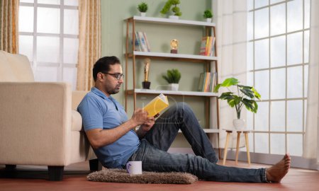 Téléchargez les photos : Plan large de l'homme indien livre de lecture en buvant du café ou du thé tout en étant assis sur le sol à la maison concept de rafraîchissement, de connaissances et de passe-temps week-end. - en image libre de droit