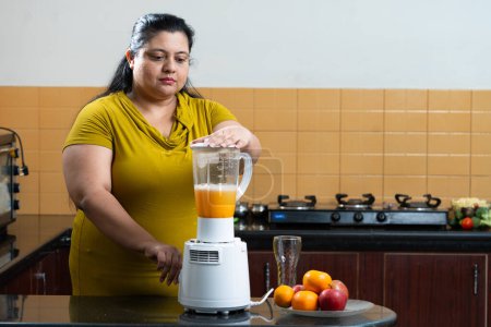 Übergewichtige indische Frau mit Mixer oder Entsafter Mixer Mühle für die Herstellung von gesundem Fruchtsaft in der Küche. Konzept für gesunde Ernährung. Kopierraum