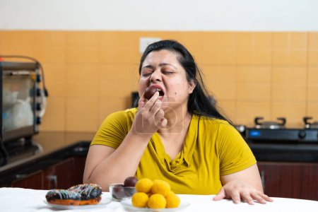Übergewichtige indische Frau, die Gulab Jamun und Donuts, Laddu oder Dessert isst, stellt sich auf einen Tisch in der Küche. Ungesunde Ernährung, Verlangen Konzept.