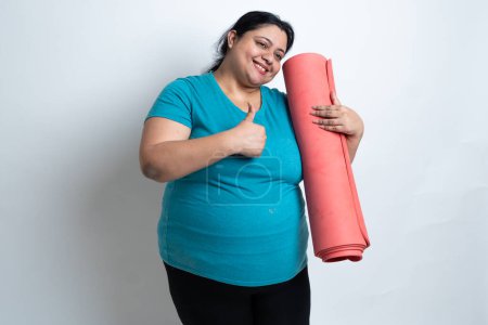 Übergewichtige, dicke indische Frau hält Yogamatte in der Hand und macht Daumen hoch. isoliert über weißem Hintergrund. Plus Size weiblich. Kopierraum