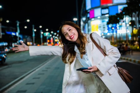 Foto de Asian business woman walking to hail waving hand taxi on road in city street at night, Beautiful woman smiling using smartphone application hailing with hand up calling cab outdoor after late work - Imagen libre de derechos
