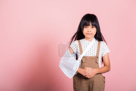 Téléchargez les photos : Arm broken. Asian little kid 10 years old hand bone broken from accident with arm splint at studio shot isolated on pink background, Happy child girl accident insurance and extreme sports speed - en image libre de droit