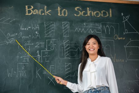 Téléchargez les photos : Back to school concept. Happy beautiful young woman standing hold pointer to back board, Asian female teacher smiling with wooden stick pointing to blackboard at school in classroom, Education - en image libre de droit