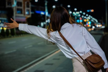 Foto de Mujer de negocios asiática granizo saludando taxi de la mano en la carretera en la calle de la ciudad por la noche, hermosa mujer sonriendo utilizando la aplicación de teléfono inteligente que llama con la mano hasta cabina al aire libre después del trabajo tardío, vista trasera - Imagen libre de derechos