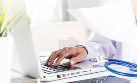 Téléchargez les photos : Médecin ou infirmière femme en uniforme avec stéthoscope elle dactylographier des informations de prescription du patient à partir de la paperasserie formulaire d'antécédents médicaux dans un ordinateur portable pour enregistrer le rapport de données de document - en image libre de droit