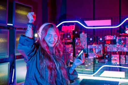 Foto de Ganando. Mujer joven sintiéndose emocionada en la sala de juegos, la gente feliz jugador jugando videojuego en línea con el teléfono móvil inteligente con luces de neón levanta las manos para ganar celebrando, sonreír con el teléfono inteligente - Imagen libre de derechos