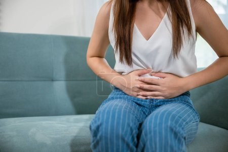 Asian young woman unhappy hands holding on stomach suffering from abdominal pain on sofa at home in living room, Sad female close eyes she painful stomachache, medical care and health concept