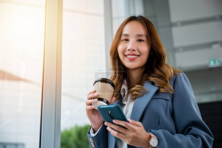 Téléchargez les photos : Jeune femme d'affaires asiatique portant une chemise bleue tenant une tasse de café et son smartphone. femme d'affaires souriant debout tenant téléphone mobile près de la fenêtre et fond flou ascenseur dans le bureau. - en image libre de droit