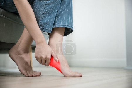 Photo for Asian beautiful female problems with foot at home, painful ankle injury, Young woman sitting on sofa holding her feet and stretch muscles have symptoms feeling pain, Health care and medical concept - Royalty Free Image