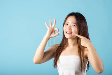 Téléchargez les photos : Concept de santé dentaire. Asiatique jeune femme pointant vers ses dents blanches fortes et montrant OK signe pour bon studio tourné isolé sur fond bleu, belle femelle pointe les doigts vers la bouche souriante - en image libre de droit