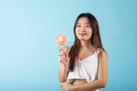 Photo for Asian beauty young woman holding pink portable electric mini fan near her face studio shot isolated on blue background, Female hand hold small plastic fan handheld she enjoying cool wind blowing - Royalty Free Image