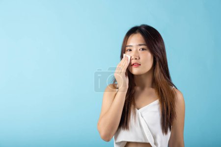 Téléchargez les photos : Facial Beauty Treatment. Asian beautiful young woman removing cosmetic make up using pad cotton face cleaner disc, studio shot isolated on blue background, Portrait of happy female cleaning face - en image libre de droit
