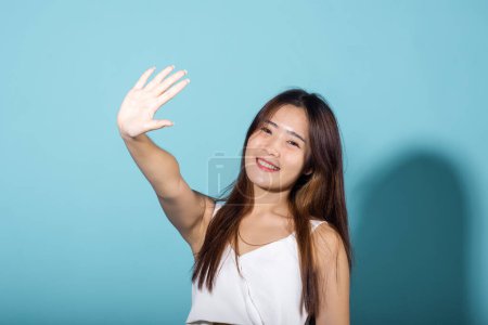Foto de Mujer levantando el brazo y la mano en la protección y bloquear el sol en el estudio de tiro aislado sobre fondo azul, mujer joven asiática sonrisa mano hasta cubrir la cara proteger la luz del sol y UV, crema solar anunciar - Imagen libre de derechos
