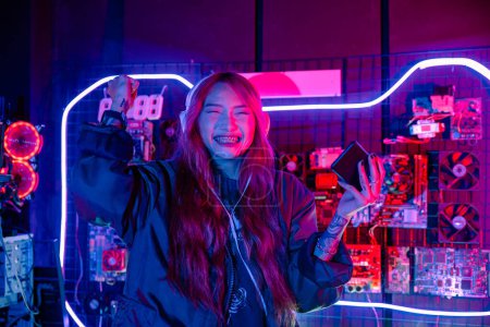 Foto de Ganando. Mujer asiática confusa que usa auriculares para juegos sintiéndose emocionada en la sala de juegos, Happy Gamer jugando videojuegos en línea con un teléfono móvil inteligente con luces de neón levanta las manos para ganar celebrando - Imagen libre de derechos