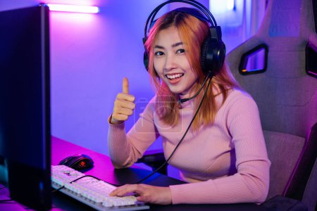 Photo for Gamer playing online game wear gaming headphones looking to camera expressing success with game giving thumbs up sign, Smiling woman live stream she play video game at home neon lights living room - Royalty Free Image