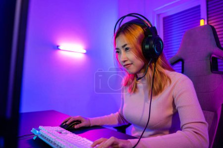 Téléchargez les photos : Happy Gamer endeavor plays online video games tournament with computer neon lights, young woman wearing gaming headphones intend to do playing live stream games online at home - en image libre de droit