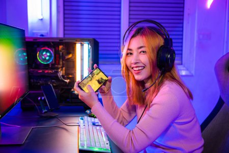 Téléchargez les photos : Gamer playing online game application on mobile phone wear gaming headphones looking to camera, Smiling young woman live stream she play video game via smartphone at home neon lights living room - en image libre de droit