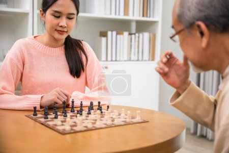 Téléchargez les photos : Beautiful young smile woman having fun sitting playing chess game with senior elderly at home, nurse caregiver in nursing home for leisure, Happy active retired people, Healthcare and medical concept - en image libre de droit