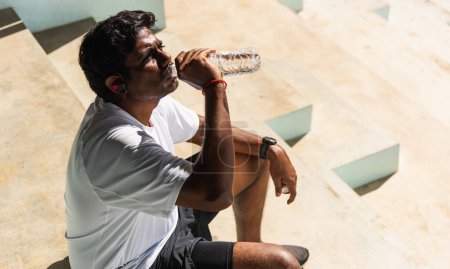 Téléchargez les photos : Gros plan Asiatique jeune coureur de sport homme noir porter des écouteurs d'athlète il boire de l'eau d'une bouteille après avoir couru au parc de santé de la rue en plein air, concept d'entraînement d'exercice sain - en image libre de droit