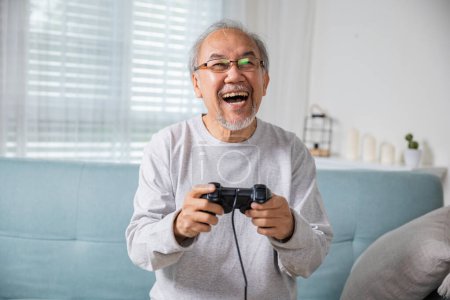 Téléchargez les photos : Asiatique senior vieil homme appréciant tenant joystick jouer à un jeu vidéo à la maison dans le salon, les mains de l'homme mature en utilisant le contrôleur de jeu, drôle sur la retraite vieux sourire assis sur le canapé vie de jeu - en image libre de droit