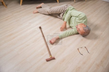 Téléchargez les photos : Malade vieillard tombant couché sur le sol parce que trébuché à la maison seul avec bâton de marche en bois dans le salon, vieillard homme grand-père ayant un accident tout en marchant avec marcheur de canne - en image libre de droit