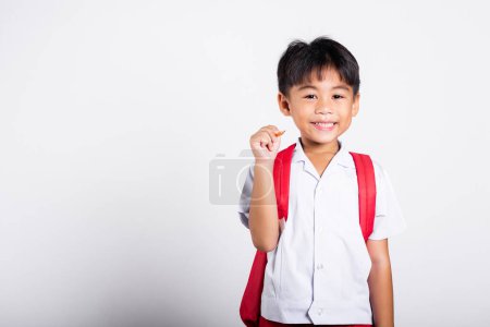 Téléchargez les photos : Asiatique tout-petit sourire heureux porter étudiant thai uniforme pantalon rouge tenant crayon pour écrivains carnet en studio tourné isolé sur fond blanc, Portrait petits enfants garçon d'âge préscolaire, Retour à l'école - en image libre de droit